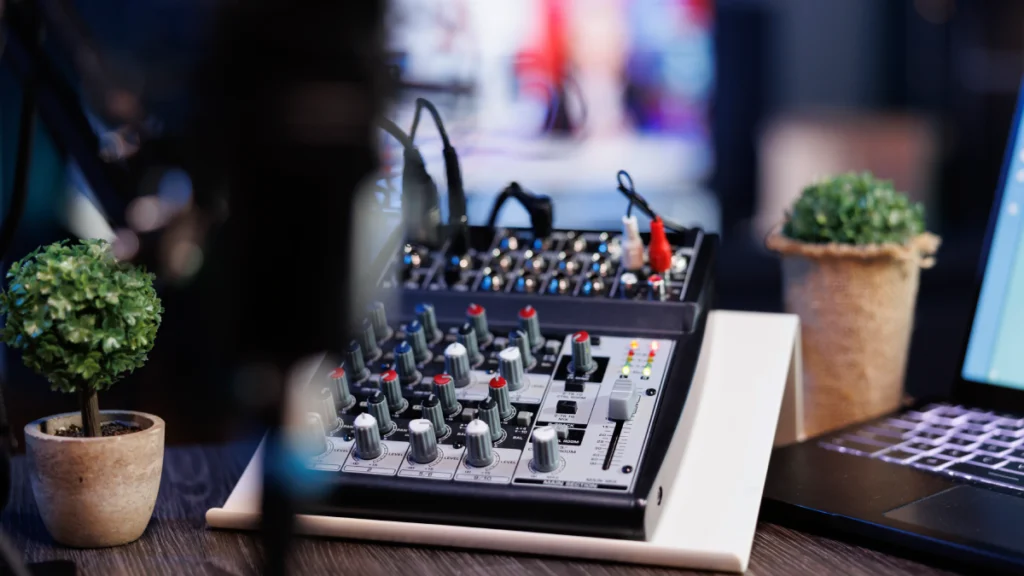 Audio mixing console with numerous knobs and faders, alongside a laptop and potted plant.