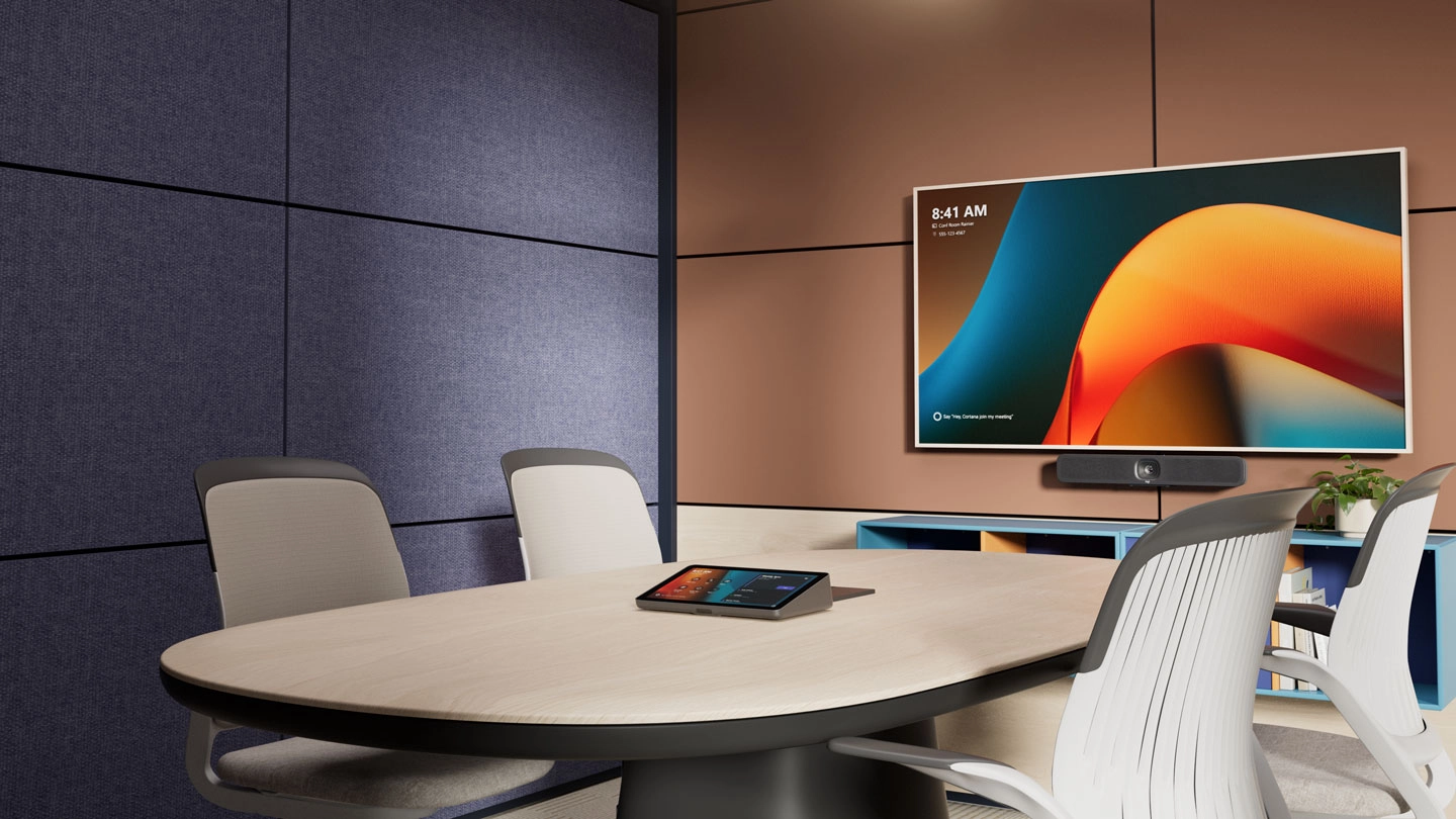 An image of a huddle room that can accommodate up to four persons. The picture shows a lone office chair, a meeting room conroller on top of a wide white working table and a large display screen on the wall.