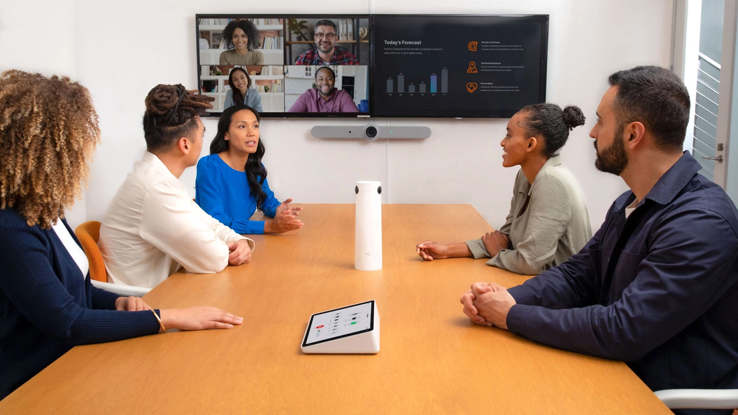 An image depicting a face to face meeting simultaneous with a remote/online co workers using dual screens, 360 degrees camera and a meeting room controller in a medium room that can accommodate up to fourteen.